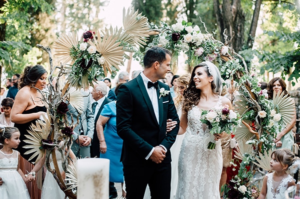 Εντυπωσιακή ξύλινη αψίδα για στολισμό δεξίωσης γάμου με αποξηραμένα φύλλα φοίνικα και μπορντό λεπτομέρειες