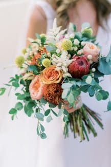 Εντυπωσιακή νυφική ανθοδέσμη με κόκκινη πρωτέα, τριαντάφυλλα και άλλα λουλούδια σε ζεστές αποχρώσεις